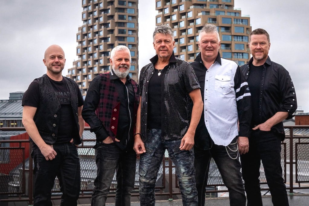 Fem män står på en takterrass med moderna byggnader i bakgrunden. De är klädda i stiliga outfits och poserar för kameran. Bilden fångar en urban atmosfär och en känsla av gemenskap.
