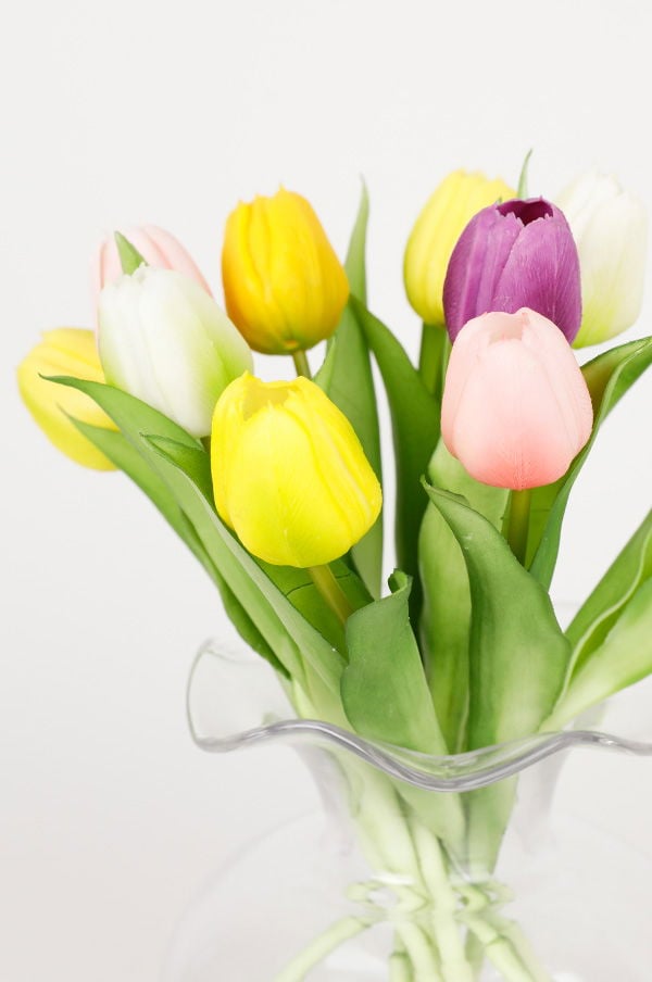 Färgglad bukett med konstgjorda tulpaner i en genomskinlig vas, inklusive gula, lila, rosa och vita blommor, mot en neutral bakgrund.