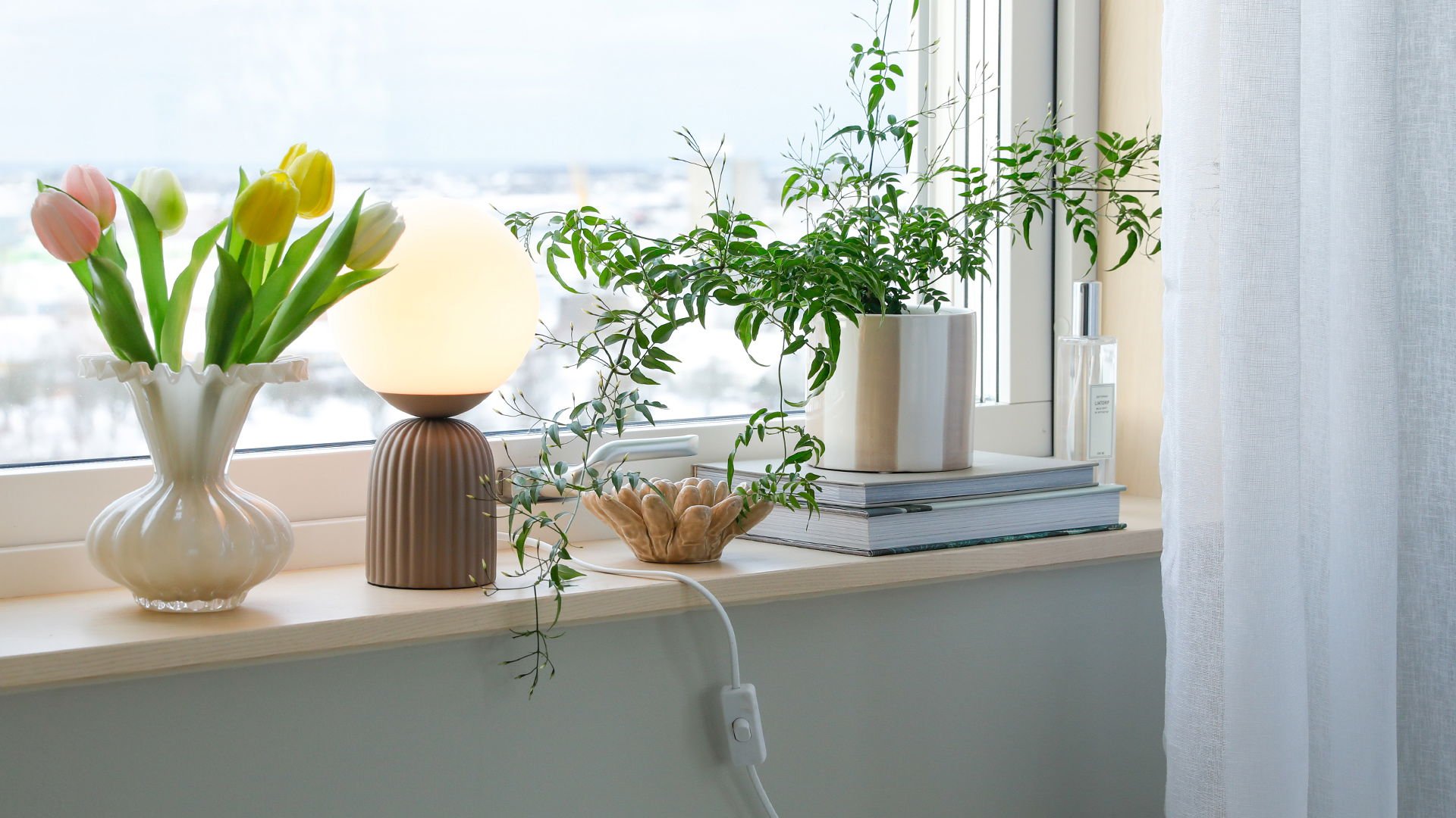 Fönsterbräda med tulpaner i en vit vas, en rund lampa, en grön växt i en kruka, och en dekorativ skål. Bakom syns böcker och en flaska med parfym. Ljusa färger och naturligt ljus skapar en lugn atmosfär.