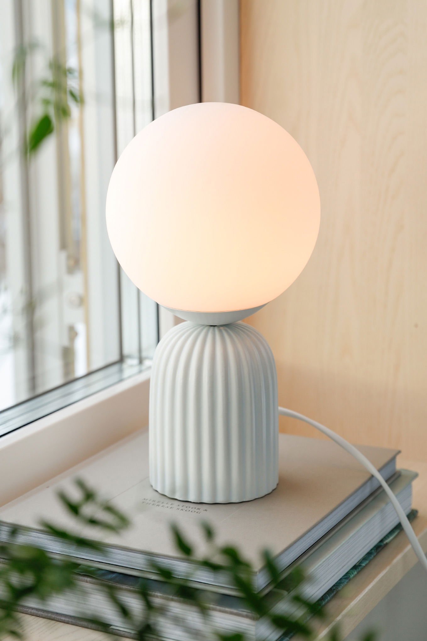 En modern bordslampa med en rund, mjuk glödlampa och en vit, ribbad bas, placerad på ett bord med böcker och nära ett fönster med växtlighet i bakgrunden.