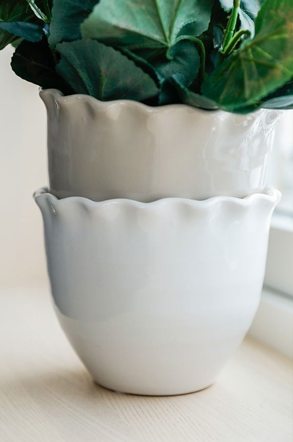 Två eleganta keramikblomkrukor i vitt med vågiga kanter, en ovanpå den andra, med grön växt ovanför, placerade på ett ljust träbord.