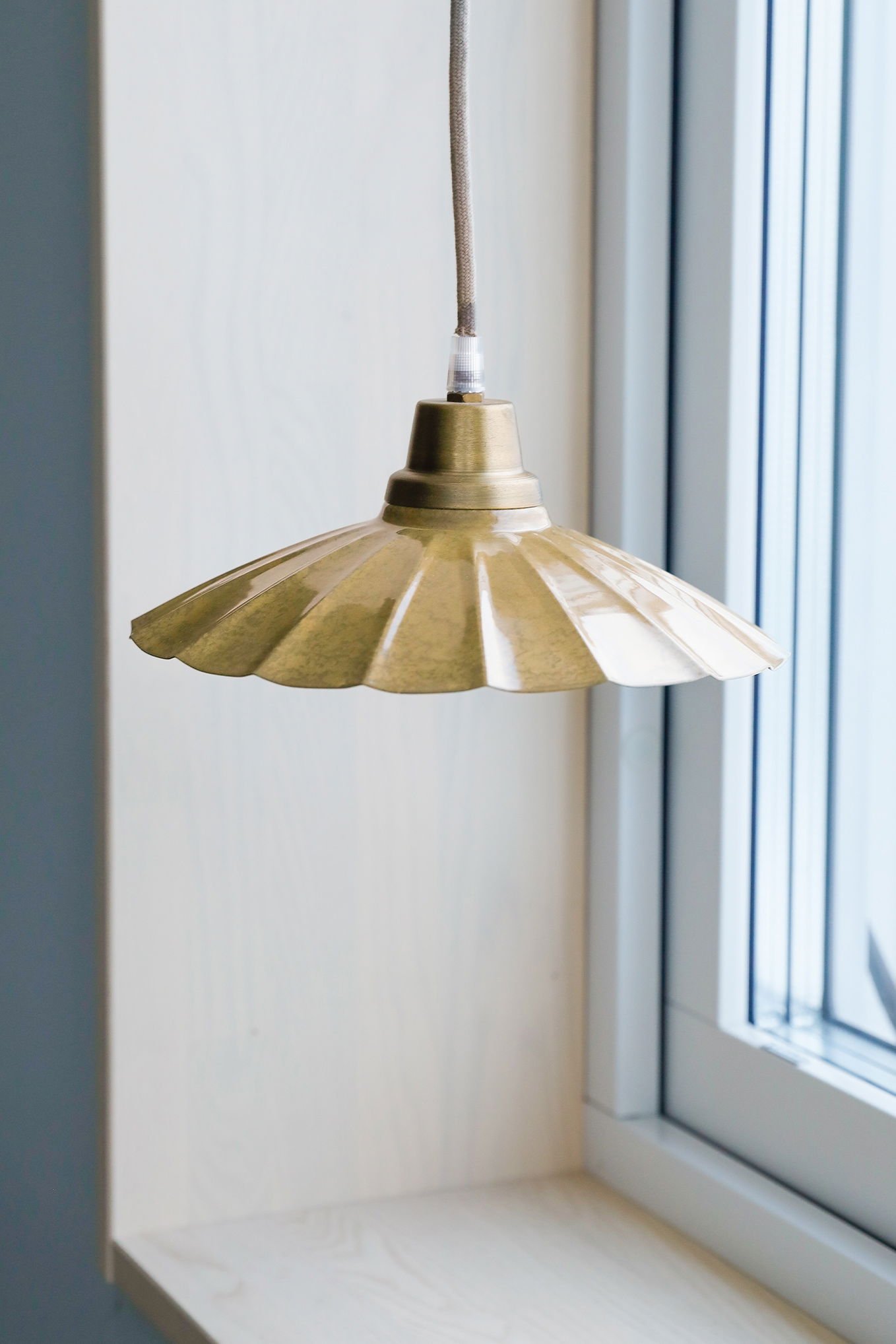 Hängande lampa i metall med en guldfärgad skärm, placerad nära ett fönster i en modern inredning.