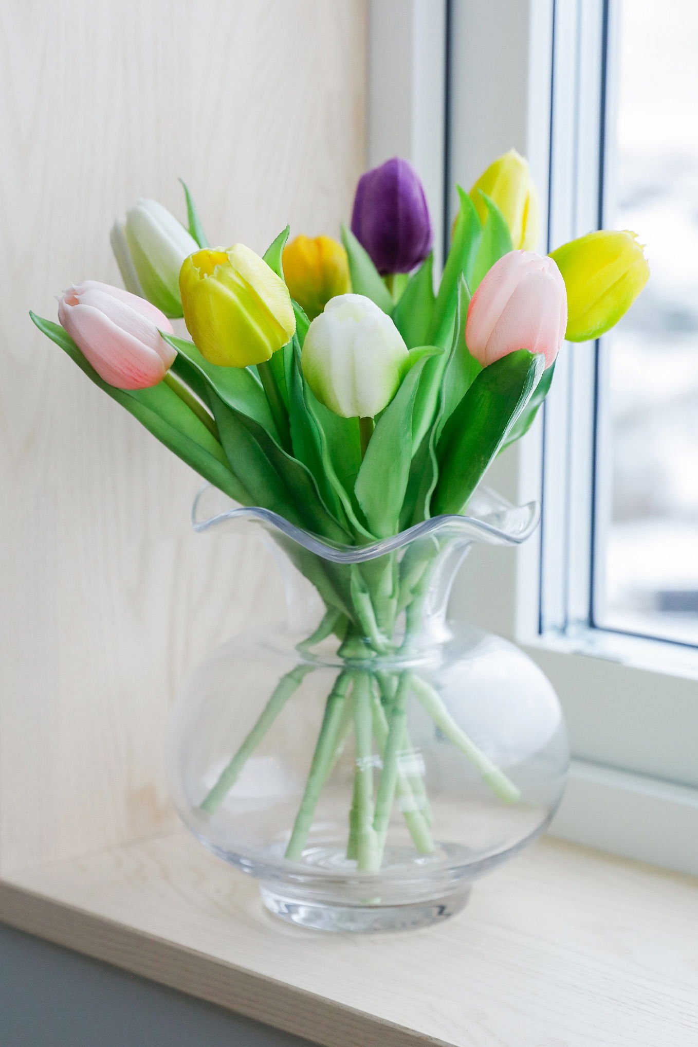 En bukett med färgglada tulpaner i en glasvas, placerad på en fönsterbräda. Tulpanerna är i nyanser av rosa, vitt, gult, lila och grönt, vilket ger en livfull och fräsch känsla i rummet.