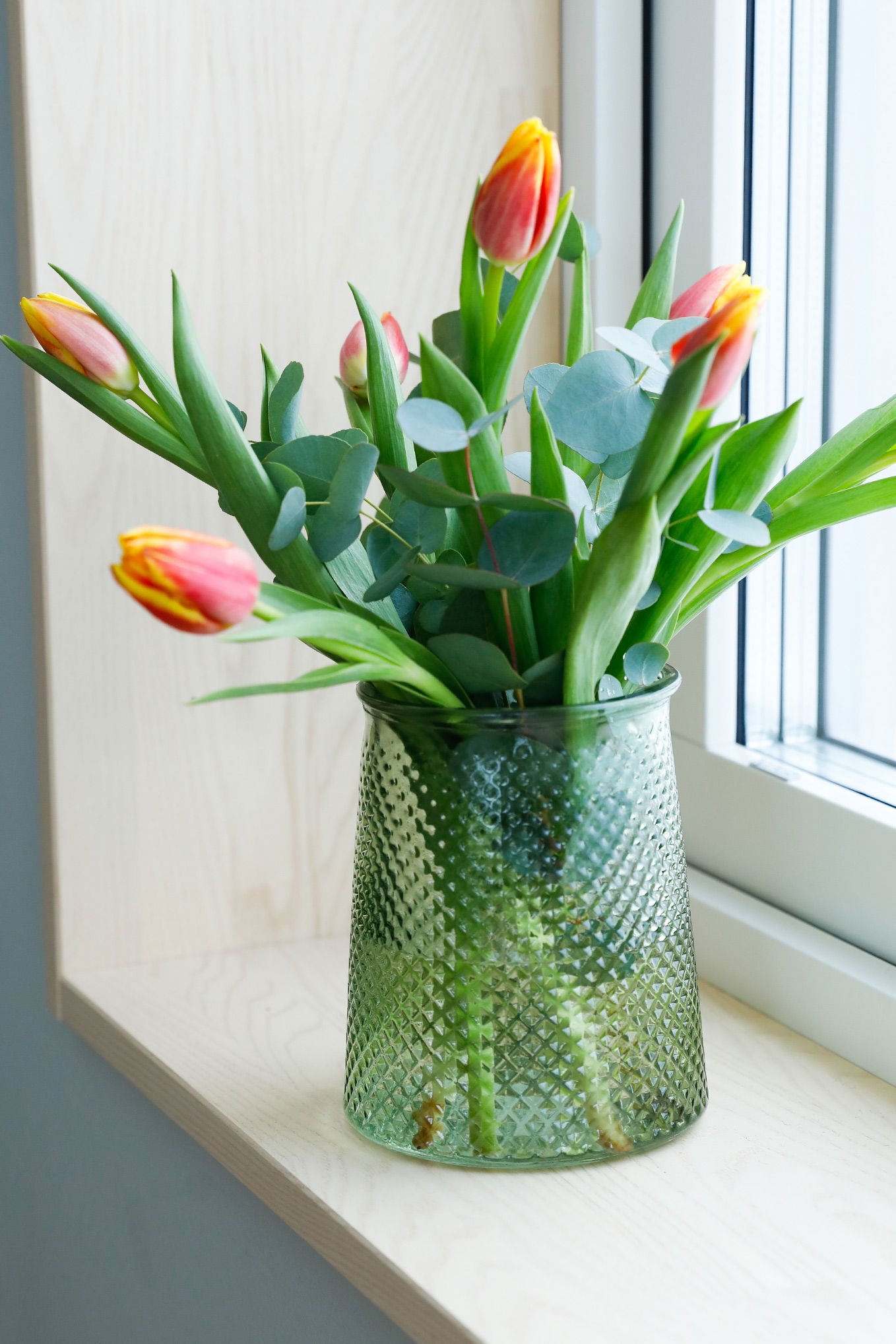Blommor i en grön glasvas med tulpaner och eukalyptus, placerade på en fönsterbräda i ett ljust rum.