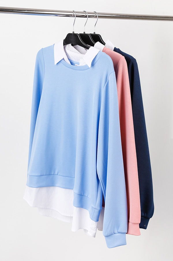 Tre färgglada sweatshirts hängande på en klädhängare, inklusive en ljusblå, en rosa och en mörkblå, med en vit skjorta som syns under. Perfekt för avslappnade outfits och säsongsanpassad stil.