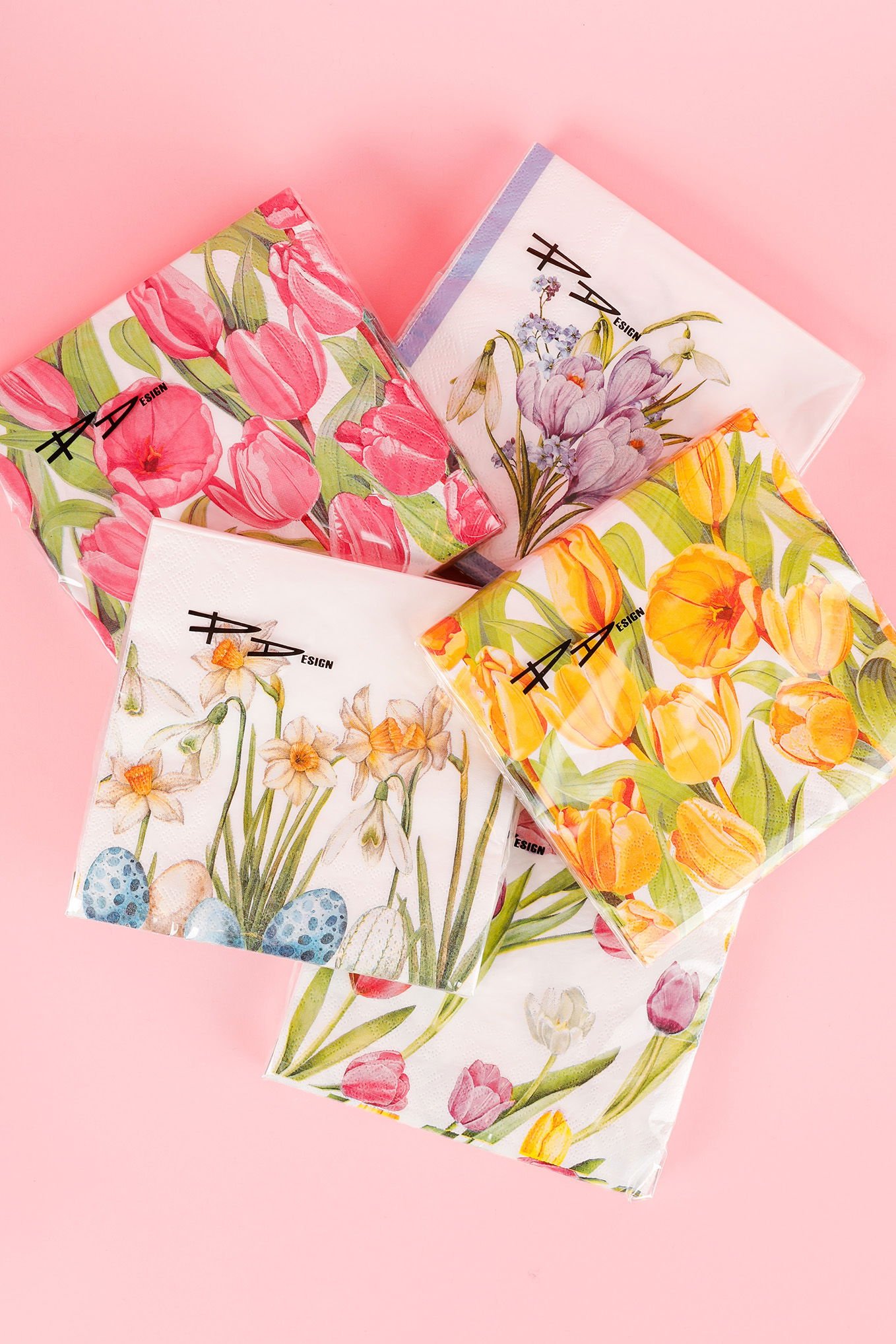 Flera färgglada servetter med blommönster, inklusive tulpaner och påskägg, på en rosa bakgrund. Perfekta för festlig dekoration och vårevenemang.