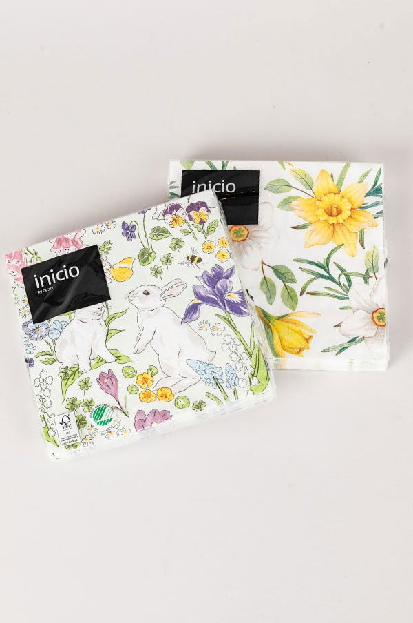 Två paket med servetter från Inicio, en med påskkaniner och blommor i färgglada mönster, och en annan med gula påskliljor och gröna blad på vit bakgrund. Perfekta för vår- och påskdekorationer.