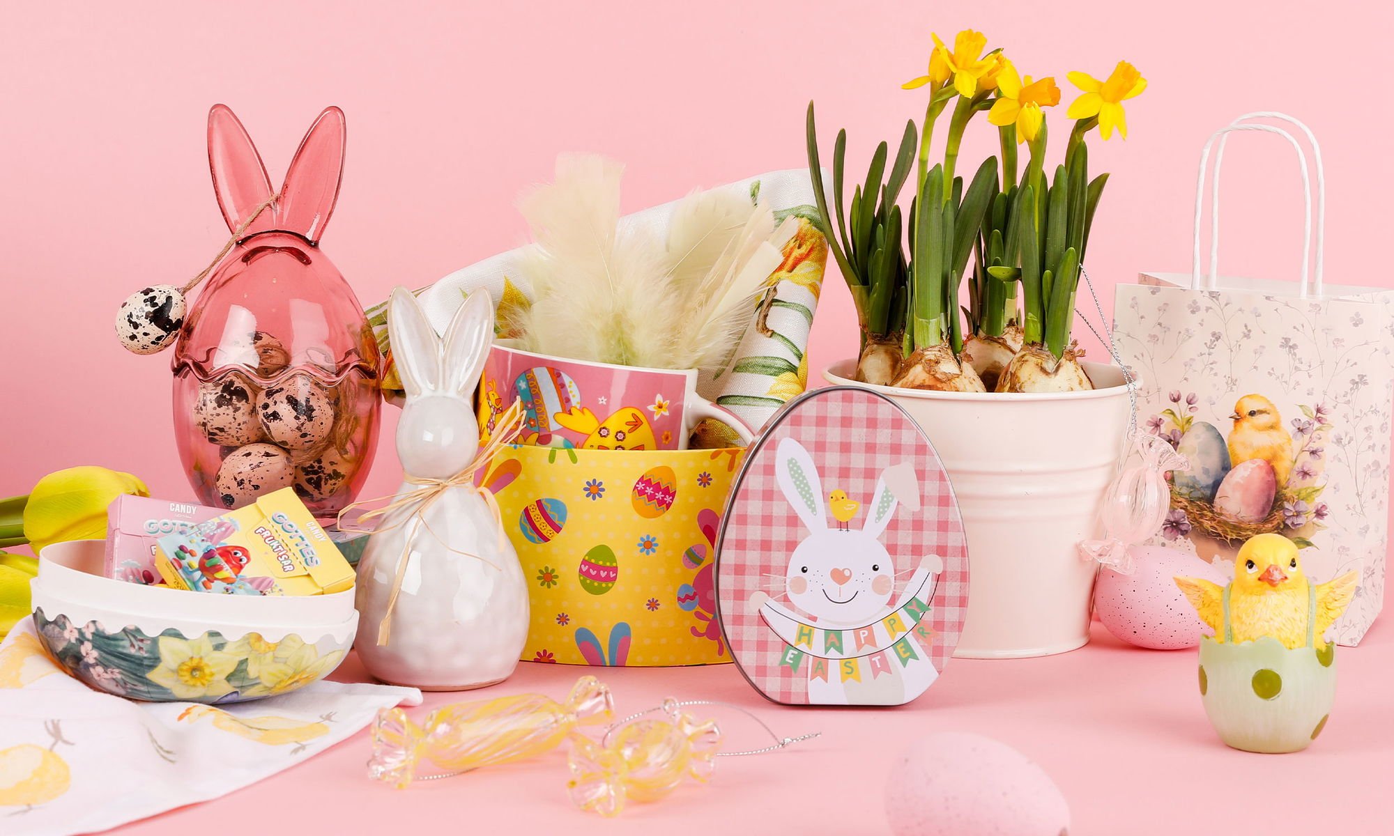 Färgglad påskdekoration med påskägg, kaninfigurer och påskliljor på en rosa bakgrund. Innehåller godis, en presentpåse och en mugg med påskmotiv. Idealisk för påskfirande och festlig dekoration.