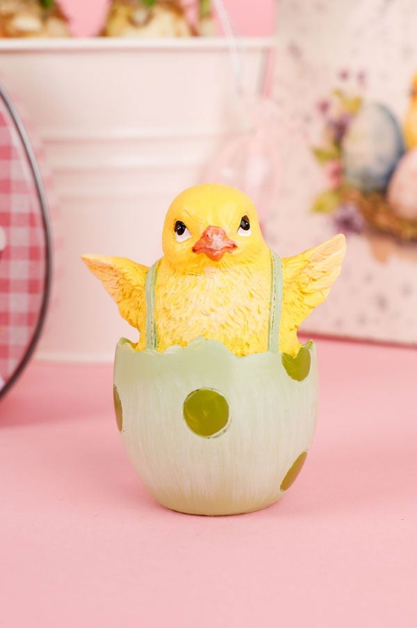 Gul kycklingfigur som sitter i ett grönt ägg med prickar, perfekt för påskdekorationer. Bakgrunden är rosa med subtila påskmotiv.
