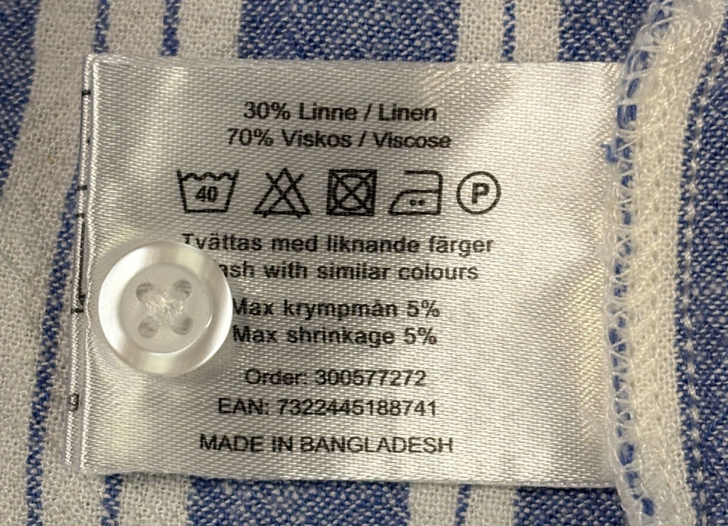 Tvättråd och materialinformation på etikett för klädesplagg, 30% linne och 70% viskos, max krympning 5%, tvättas med liknande färger, tillverkad i Bangladesh.