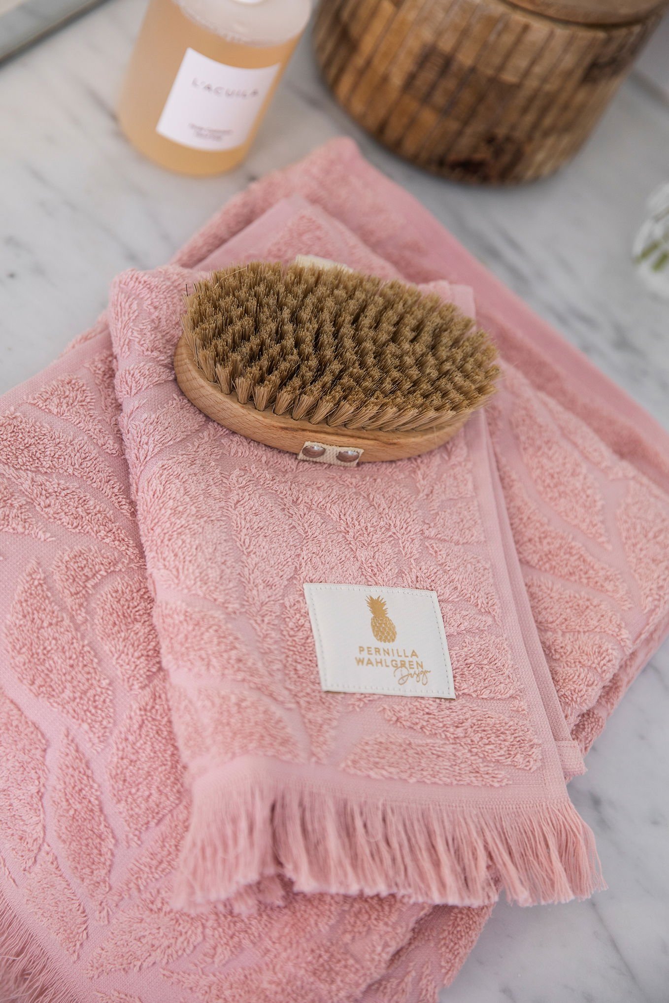 Rosa handdukar i mjuk frotté med bladmotiv, placerade på en marmor yta tillsammans med en träborste och en flaska kroppstvål. Designad av Pernilla Wahlgren.