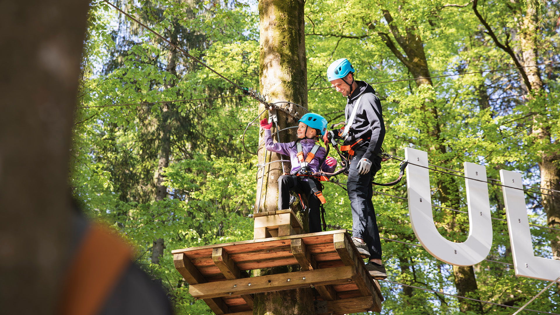 En far och dotter förbereder sig för att åka zipline i en äventyrspark, omgivna av grönskande träd. Båda bär hjälmar och säkerhetsutrustning, och dottern ser glad ut när hon håller i sig.