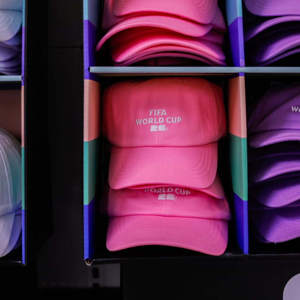 Flera färgglada kepsar i en butik, med fokus på en rosa keps som har texten "FIFA WORLD CUP" på framsidan.