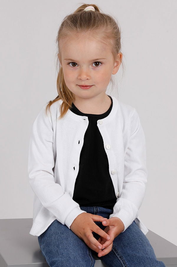 Liten flicka med långt blont hår, iförd en vit kofta och svart t-shirt, sitter med händerna i knät och ser rakt in i kameran.