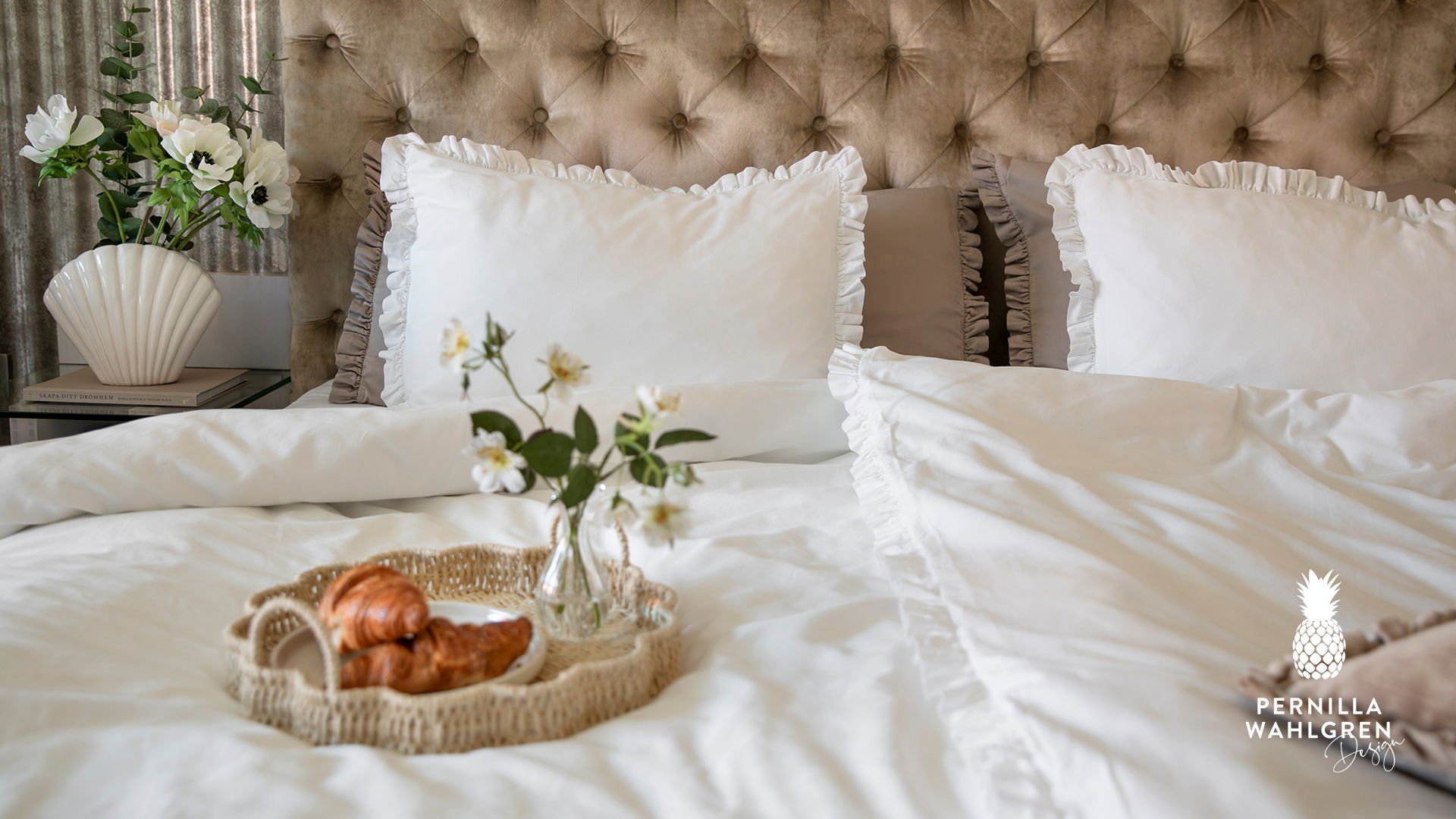 Elegant säng med vita sängkläder och prydliga kuddar med volangkant. En liten vas med blommor och en korg med croissanter på sängbordet, vilket skapar en inbjudande atmosfär.