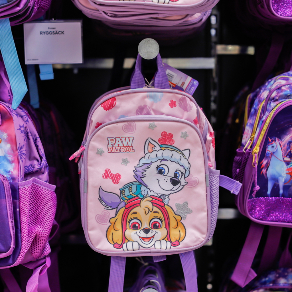 Rosa ryggsäck med Paw Patrol-tema, prydd med karaktärerna Skye och Everest, hängande bland andra väskor i en butik.