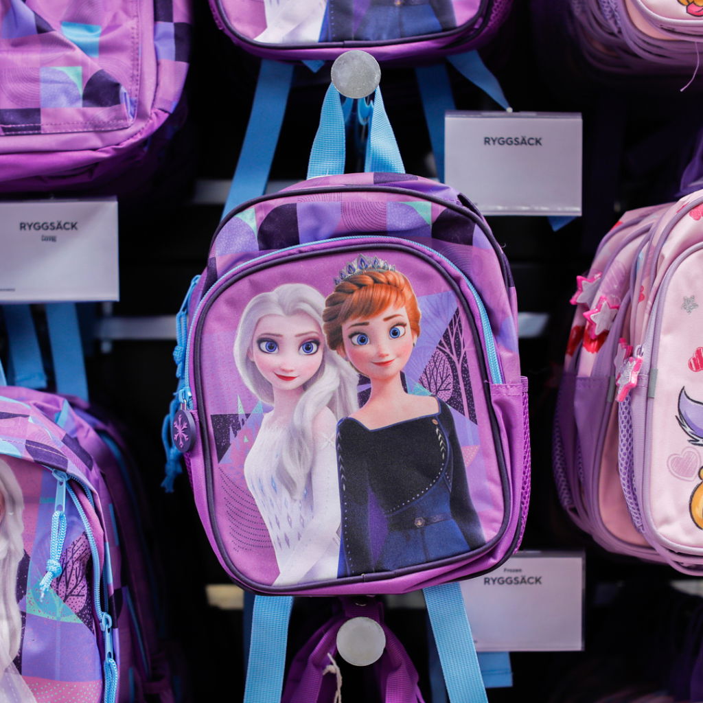 Ryggsäck med motiv av karaktärerna Elsa och Anna från filmen Frost, i lila färg med blå detaljer, hängande på en hylla.