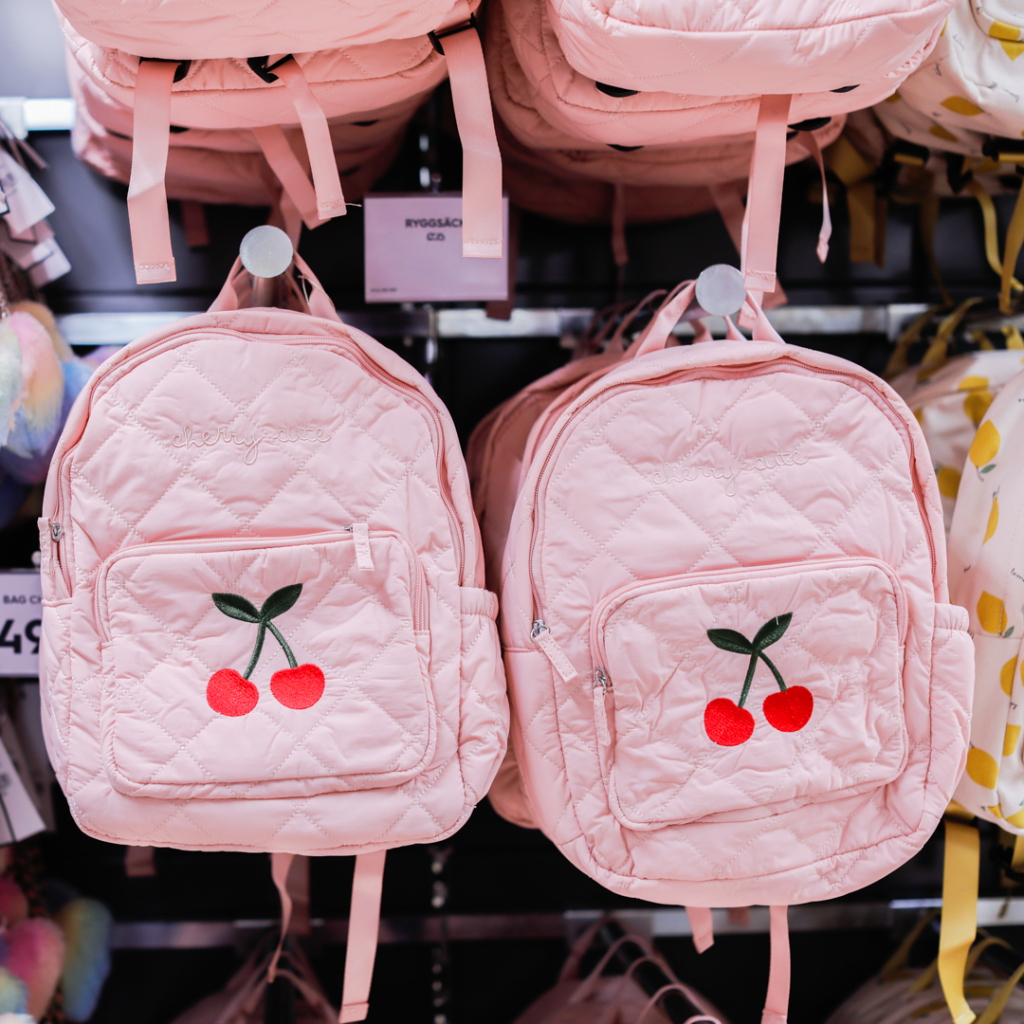 Rosa ryggsäckar med körsbärsbroderi, hängande på en klädhängare i en butik.