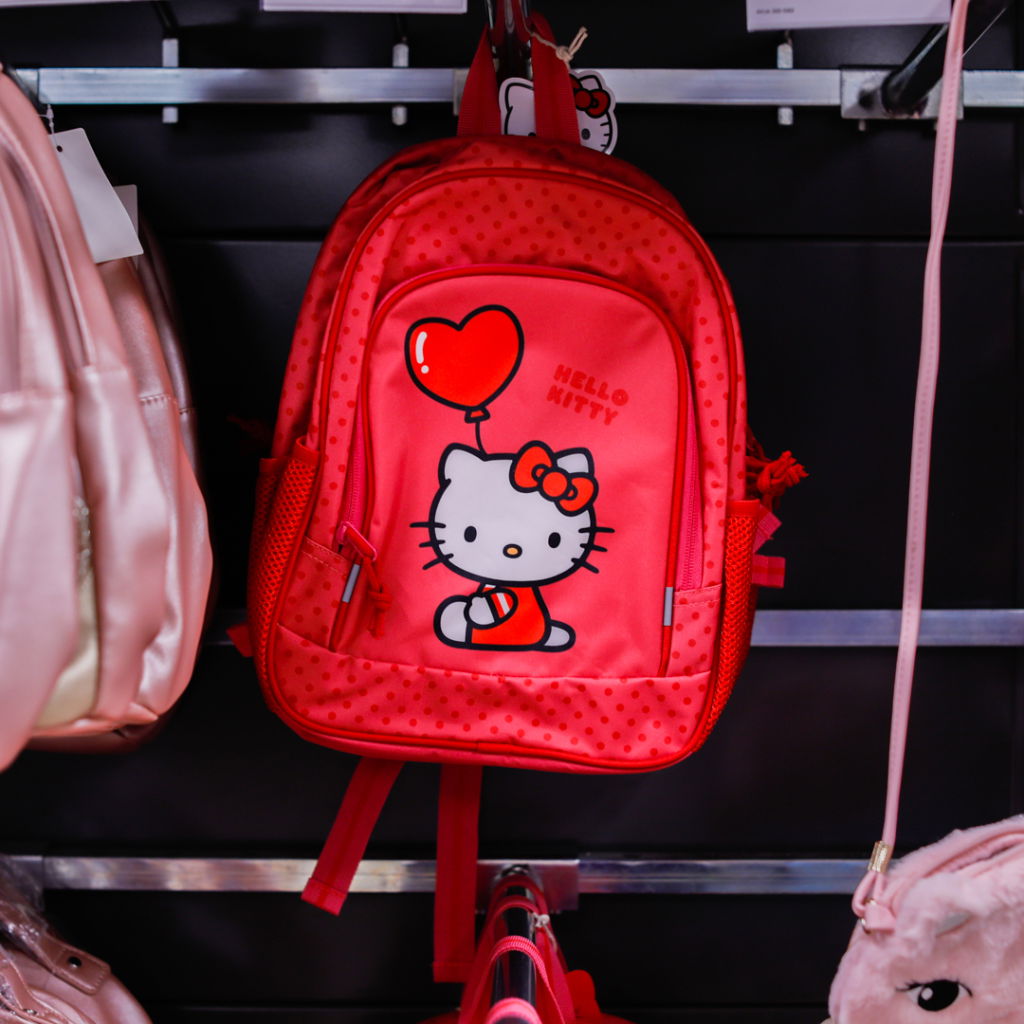 Röd Hello Kitty ryggsäck med hjärta och prickigt mönster, hängande på en hylla bland andra väskor.