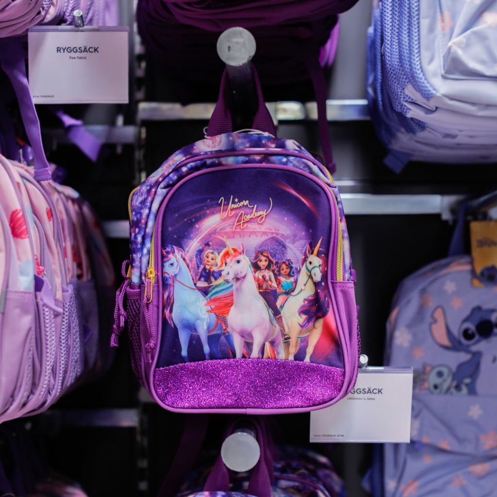 Ryggsäck med motiv från Unicorn Academy, prydd med unicorns och färgglada karaktärer, hängande på en hylla bland andra väskor.