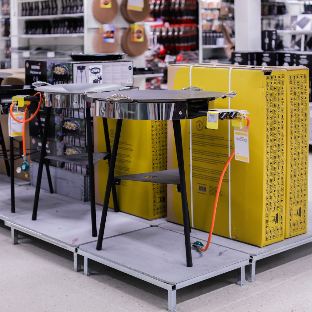 Gasolgrill med svart bord och hylla, placerad i en butik med förpackningar i bakgrunden. Grillen har en orange slang för gasanslutning och är del av ett sortiment av grillprodukter.