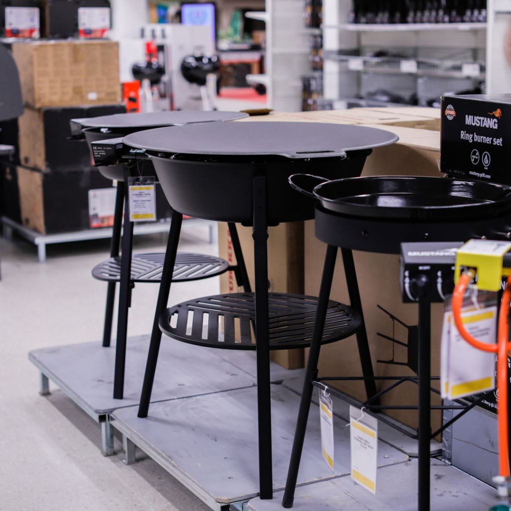 Svart gasolgrill med tre ben och hylla i en butik, omgiven av andra grillar och produkter. Perfekt för utomhusmatlagning och grillfester.
