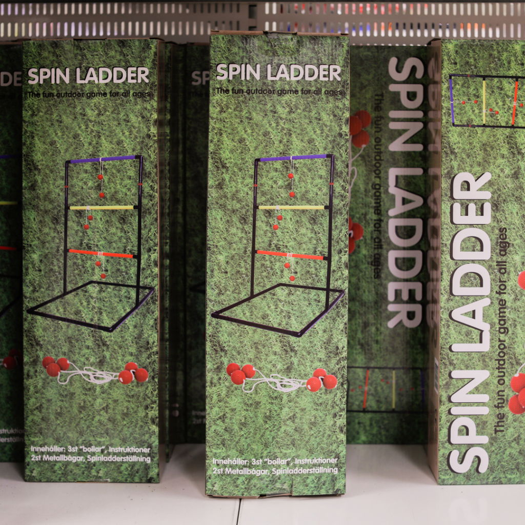 Förpackning av spelet Spin Ladder, ett utomhusspel för alla åldrar. Innehåller tre bollar, instruktioner och en metallställning. Bakgrunden visar gräs, vilket ger en lekfull känsla.
