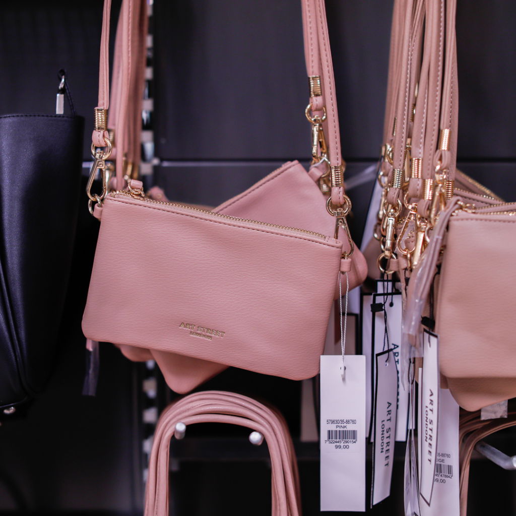 Rosa handväska i konstläder med guldfärgade detaljer, hängande på en klädhängare i butik.