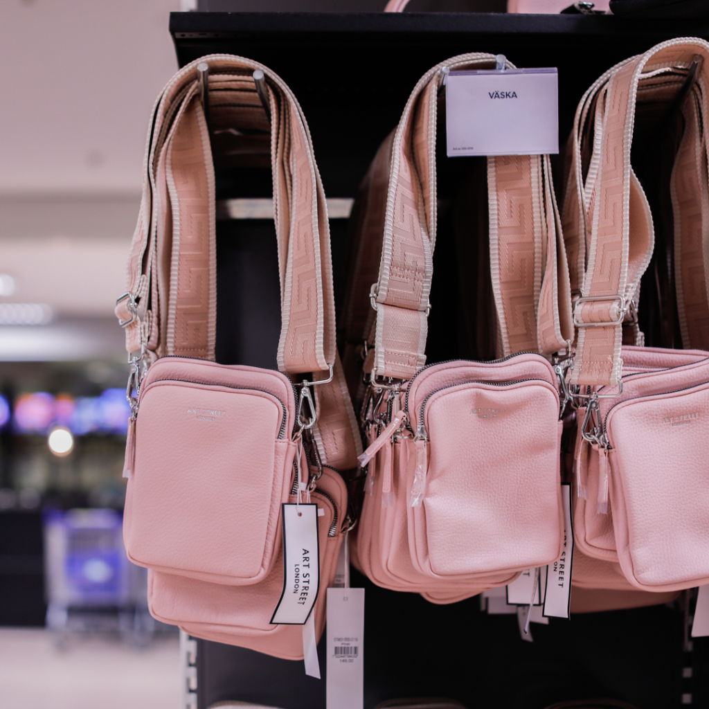 Rosa axelväskor hänger på en hylla i en butik, med märkning som visar produktinformation. Väskorna har detaljerade remmar och en stilren design, perfekta för modeintresserade.