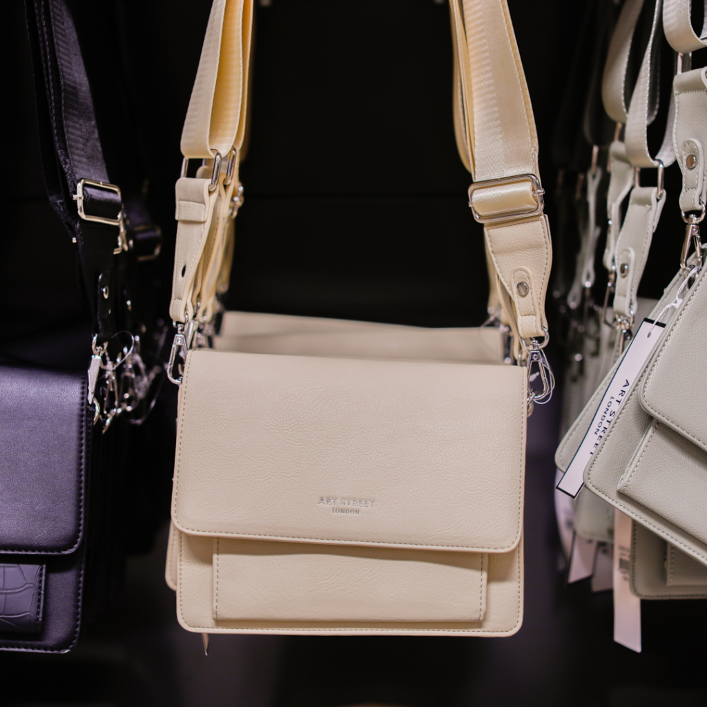 Korsningsväska i beige läder med justerbar axelrem, hängande bland andra väskor i en butik.