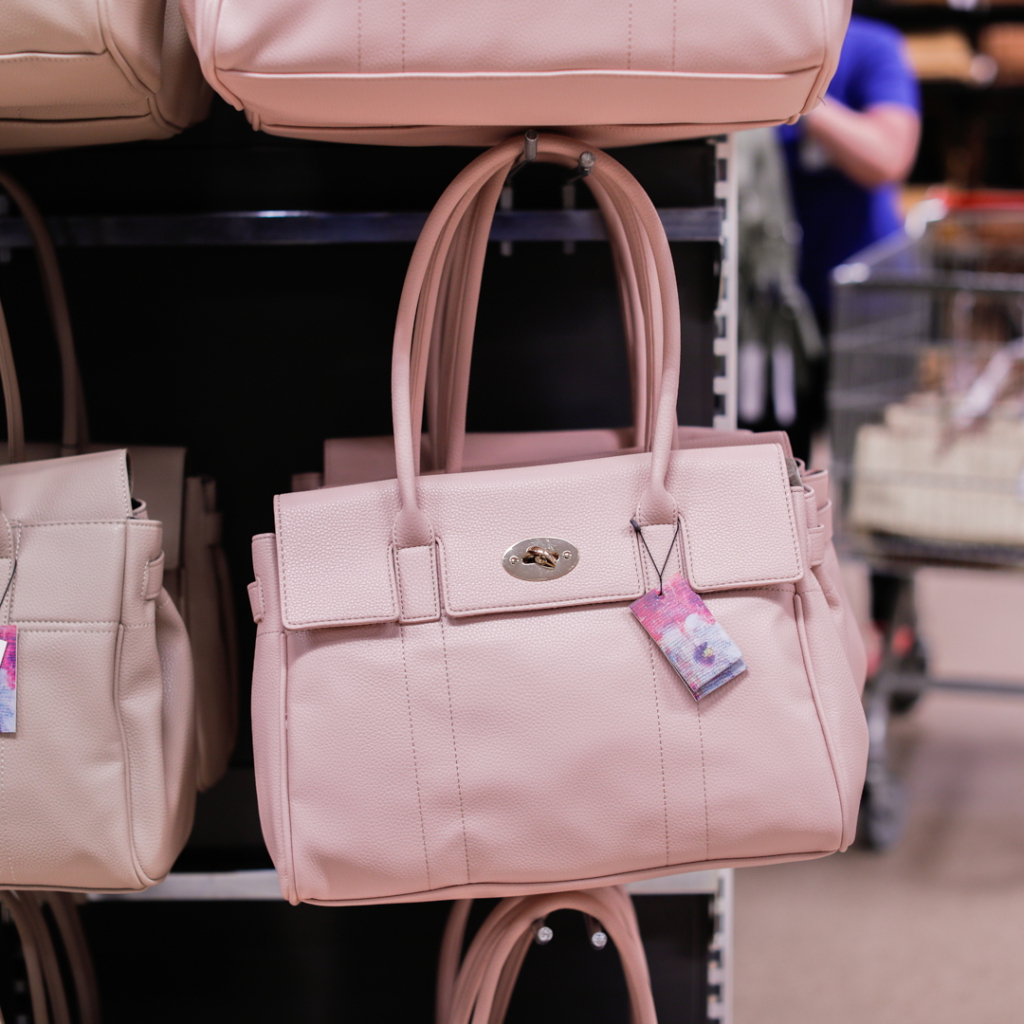 En rosa handväska i konstläder hänger på en klädhängare i en butik, med en prislapp som hänger från handtaget. Bakgrunden visar fler väskor och en kund med en kundvagn.