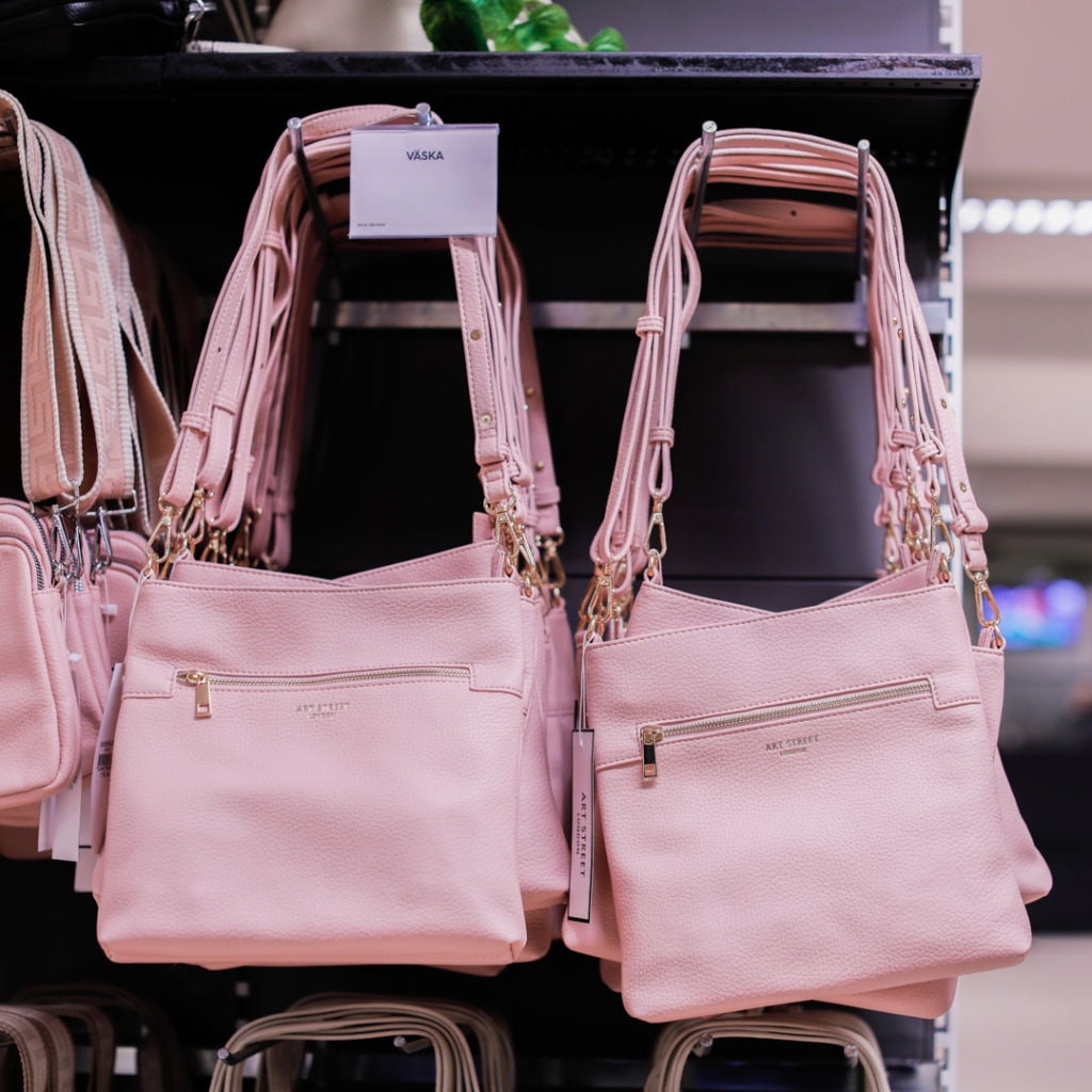 Två rosa handväskor hängande på en hylla i en butik, med en prislapp som visar ordet "VÄSKA". Väskorna har kedjor och justerbara axelremmar, vilket gör dem både stiliga och praktiska.