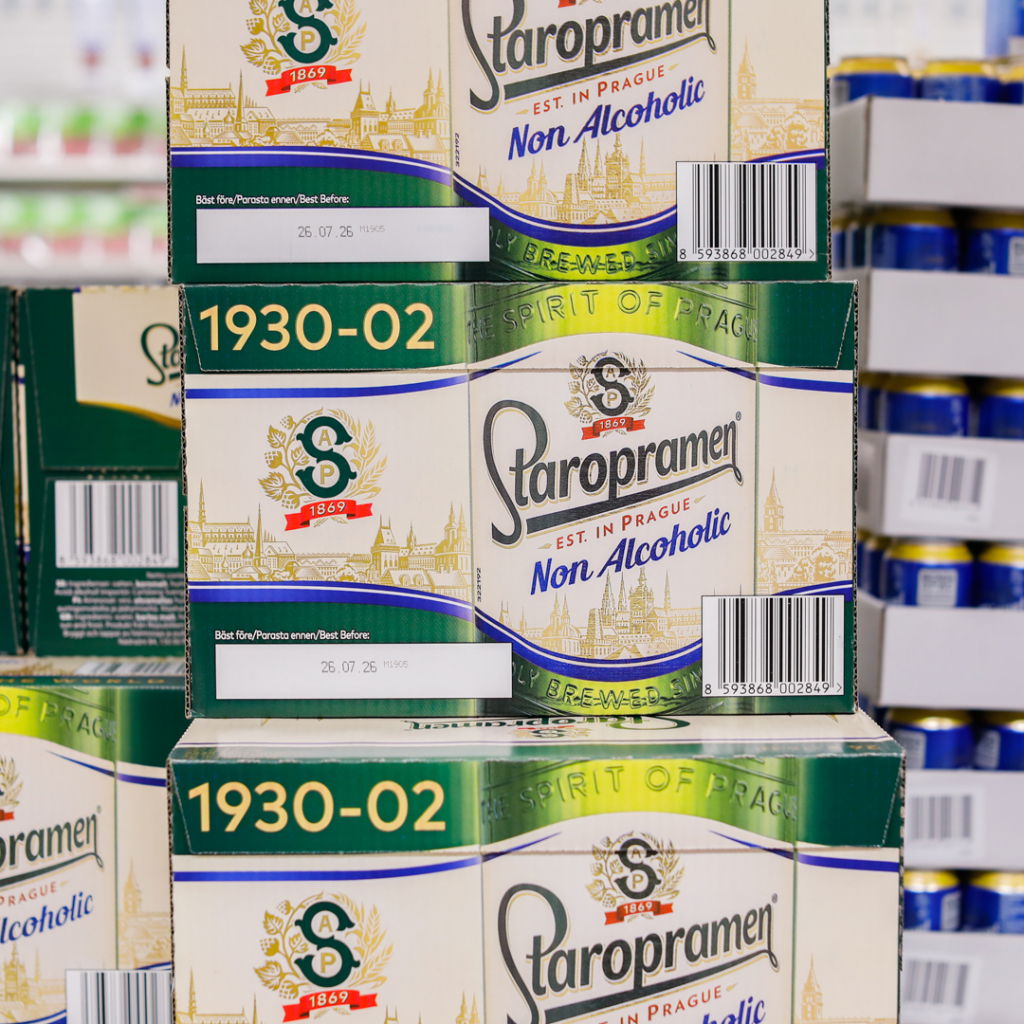 Förpackningar med Staropramen alkoholfritt öl, med tydlig märkning av produktinformation och bäst före-datum, i en livsmedelsbutik.