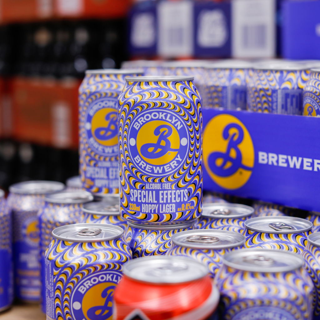 Färgglad burk av Brooklyn Brewerys alkoholfria öl "Special Effects" Hoppy Lager, 330 ml, med iögonfallande design och detaljerad etikett. Burkarna är staplade i en butiksmiljö.