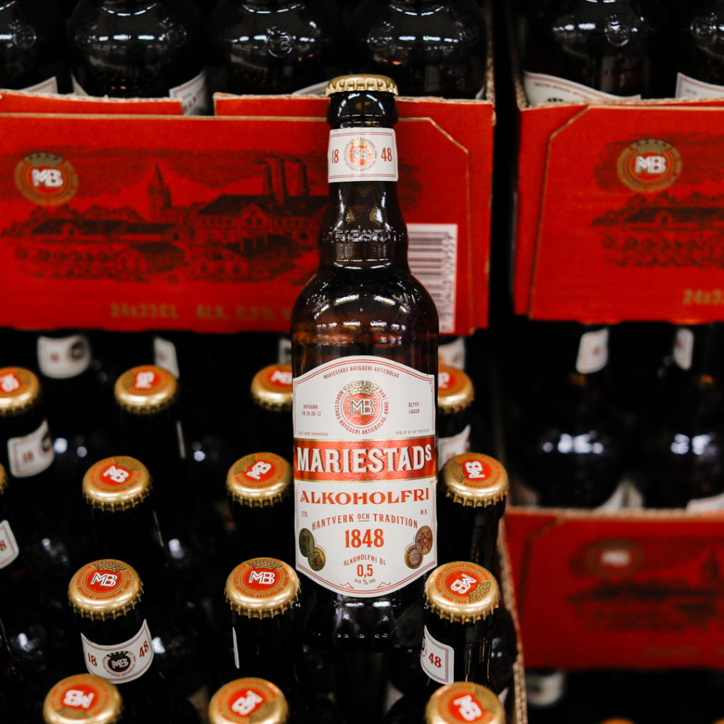 Flaska av Mariestads alkoholfri öl, 0,5% alkohol, stående framför en rad med mörka ölflaskor på en hylla. Bakgrunden visar en röd kartong med texten "Mästerbryggeriet".