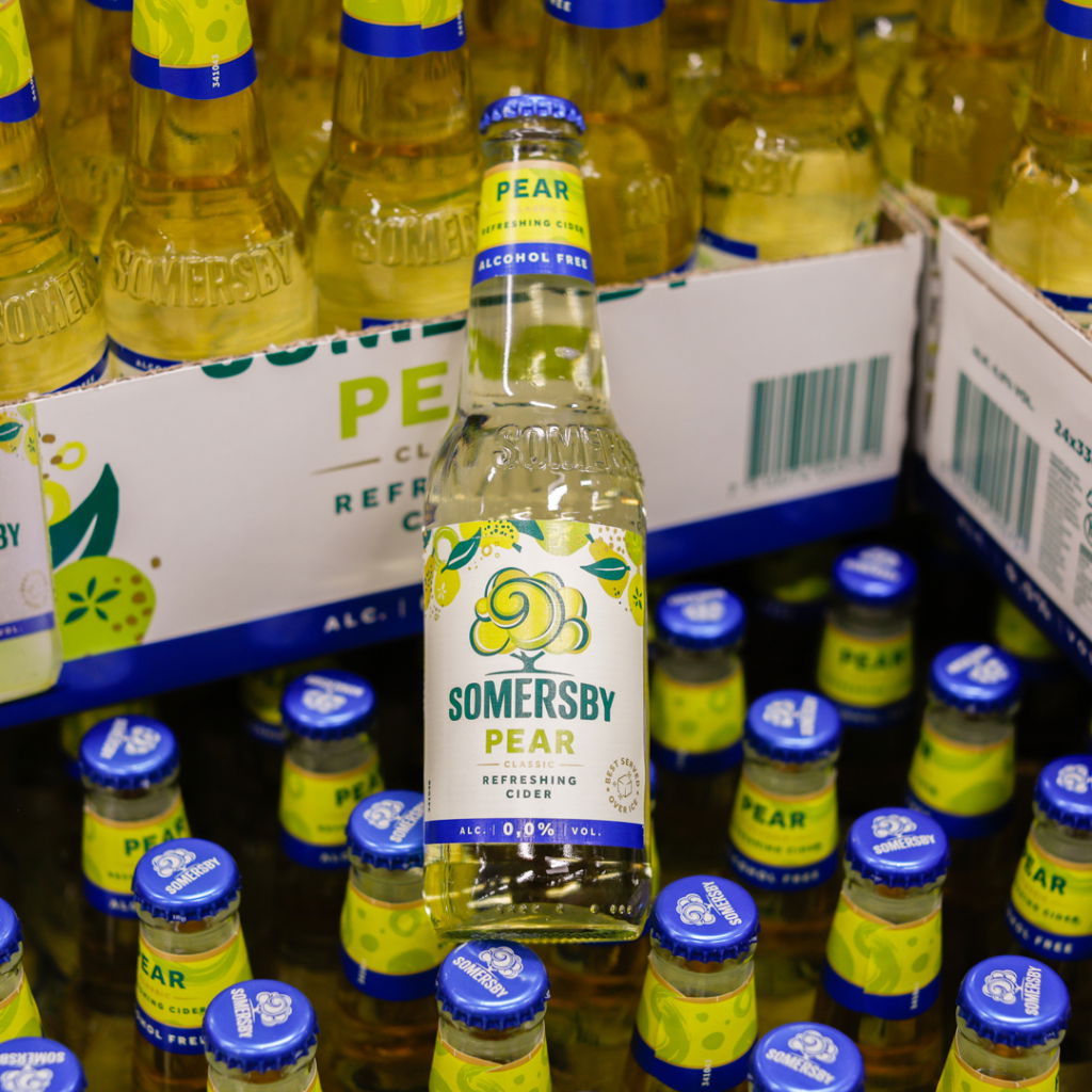 Flaska av Somersby päroncider, alkoholfri, omgiven av flera flaskor i en butik. Flaskan har en tydlig etikett med texten "Somersby Pear" och en fräsch design.