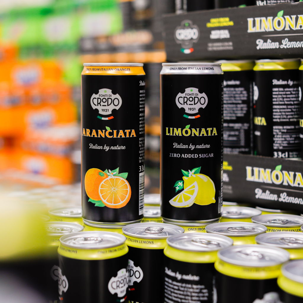 Burkar med Aranciata och Limonata från Fonti di Credo, som visar italienska citrusdrycker utan tillsatt socker. Bakgrunden har fler burkar och färgglada produkter.