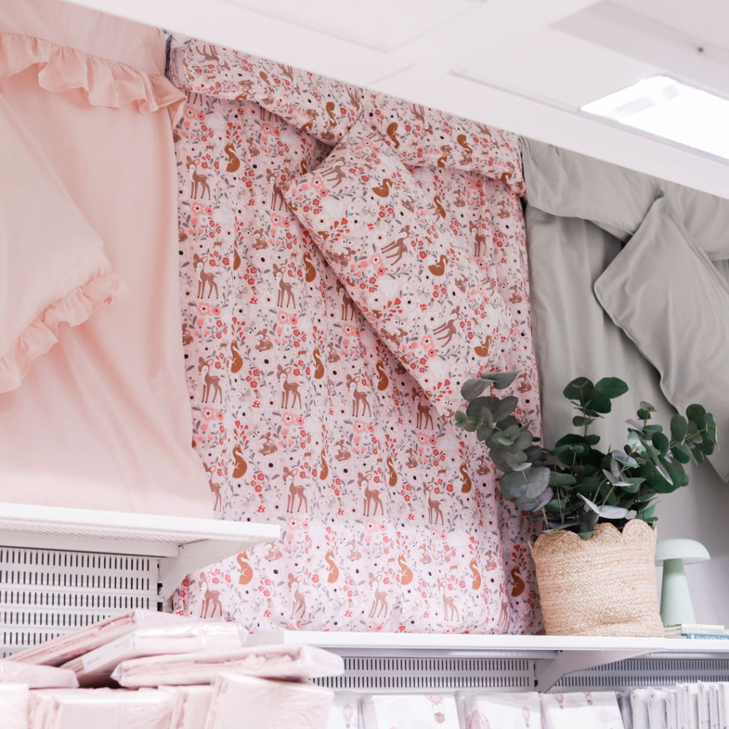 Färgglada sängkläder med djur- och blommönster i rosa och beige, tillsammans med enfärgade sängkläder i mjuk grön nyans. En dekorativ växt i korg pryder hyllan, vilket ger en hemtrevlig känsla i rummet.