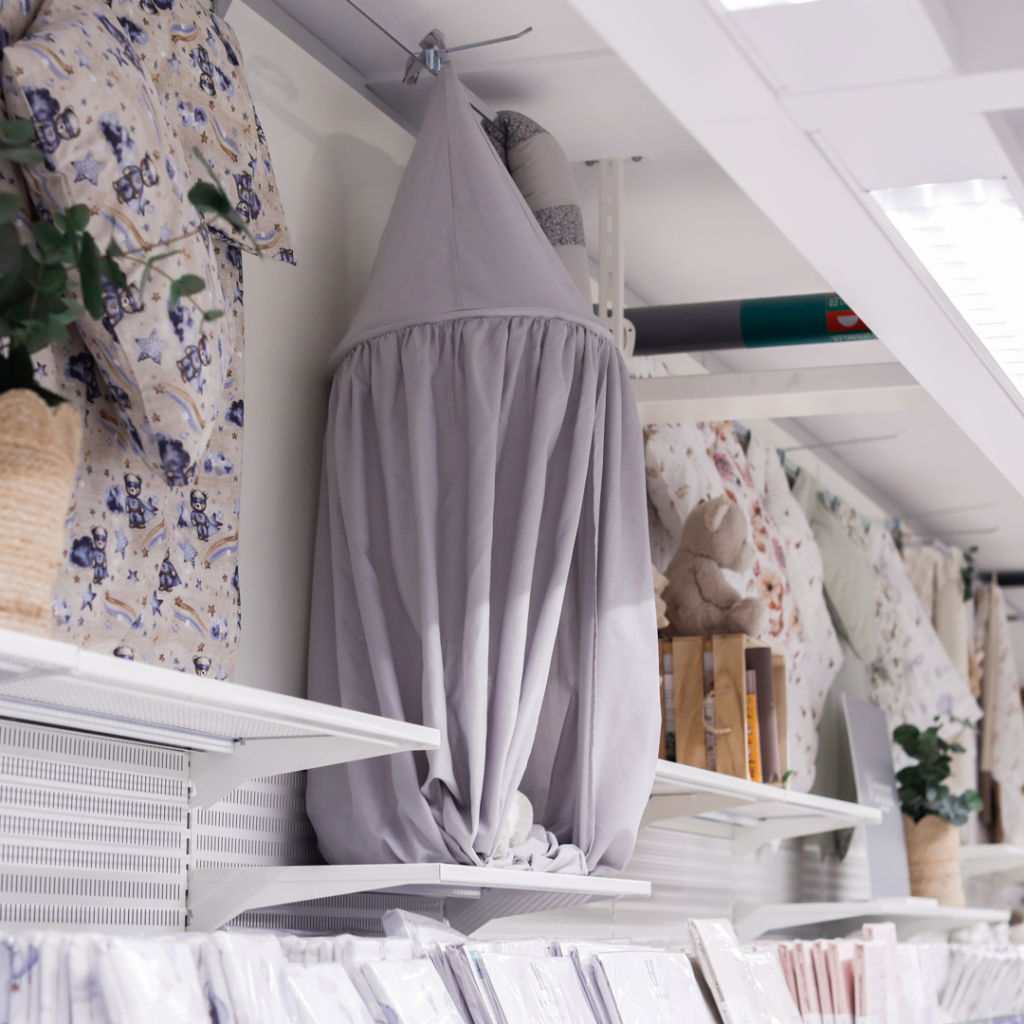 Grå hängande sänghimmel i en butik, omgiven av olika barntextilier och dekorationer på hyllorna.