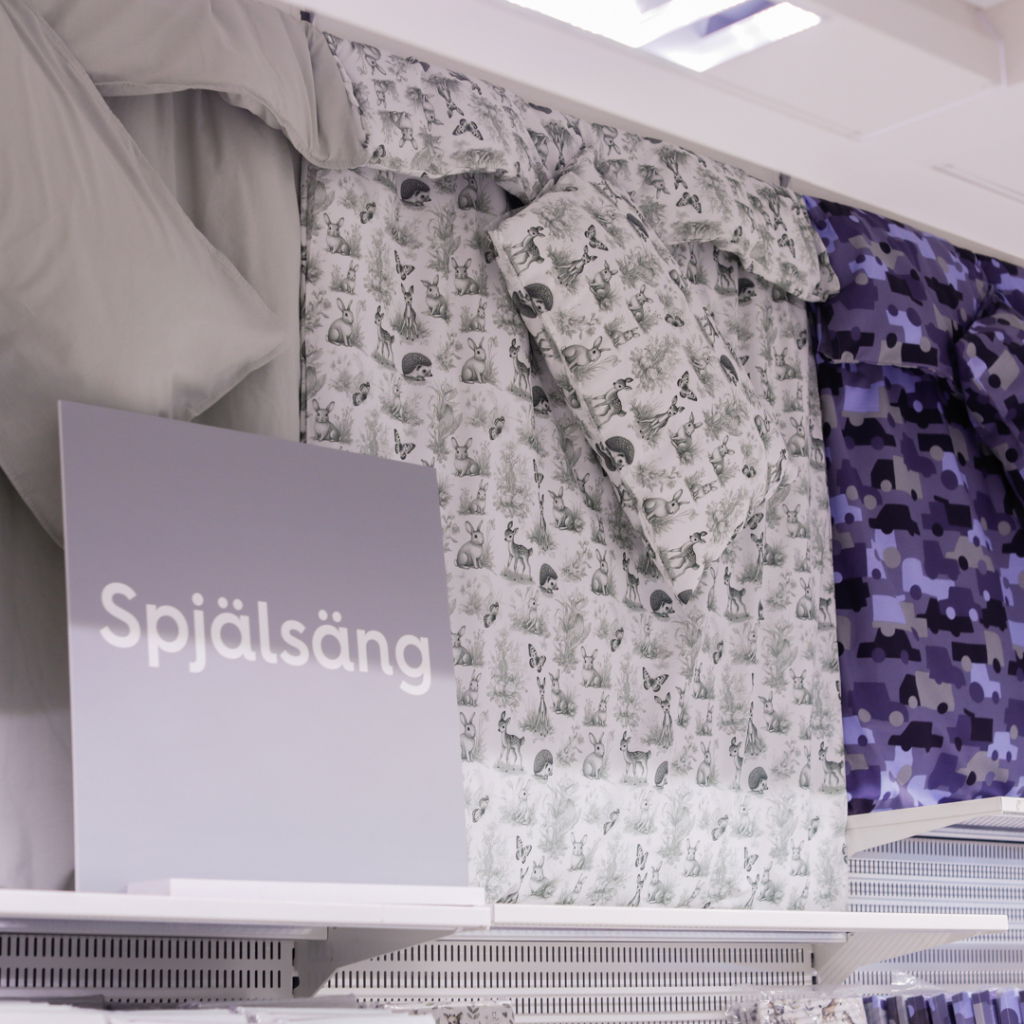 Spjälsäng med mönstrade sängkläder, inklusive ett grått set med djurmotiv och ett lila camouflagemönster, utställda i en butik.