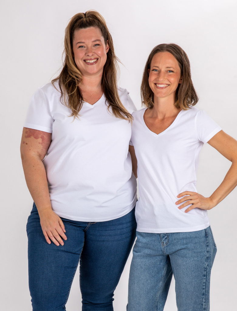 Två leende kvinnor i vita t-shirts poserar tillsammans. Den ena har längre, lockigt hår och en mer kurvig kroppstyp, medan den andra har kortare, rakt hår och en smalare kroppstyp. Båda bär jeans och står med armarna runt varandra i en vänskaplig pose.
