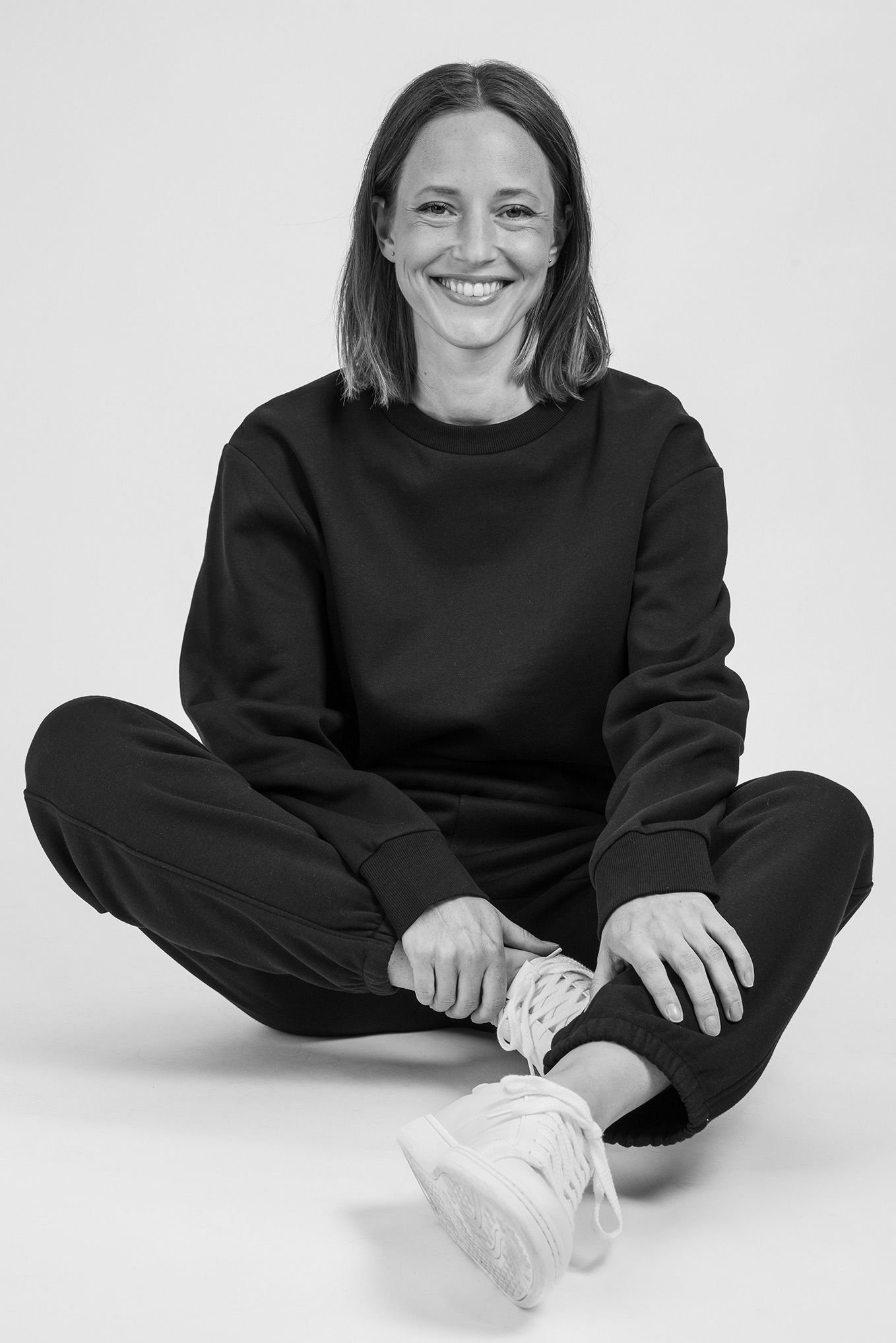Leende kvinna i svart sweatshirt och byxor, sittande på golvet med benen korsade. Hennes stil är avslappnad och modern, med vita sneakers som kompletterar outfiten. Bakgrunden är neutral och ljus.