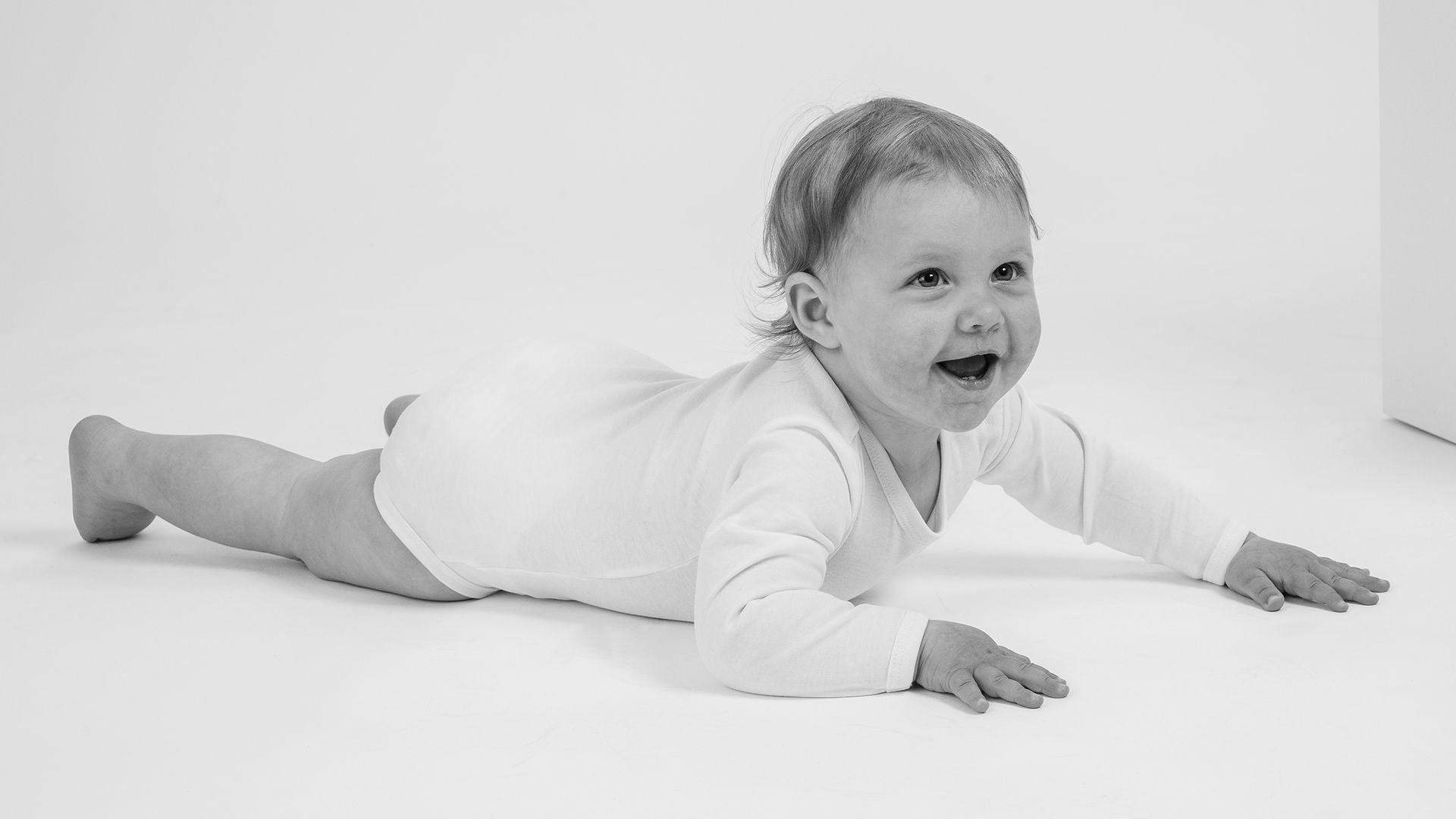 Litet barn i vit kroppströja ligger på mage och ler, på en vit bakgrund. Bilden fångar barnets glädje och lekfullhet, perfekt för att illustrera barngarderob eller babyprodukter.