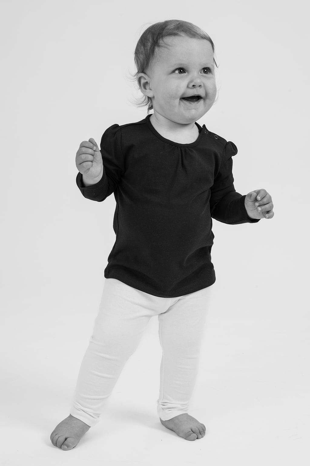 Liten glad flicka som står upp och ler, klädd i en svart topp och vita leggings, med en neutral bakgrund.
