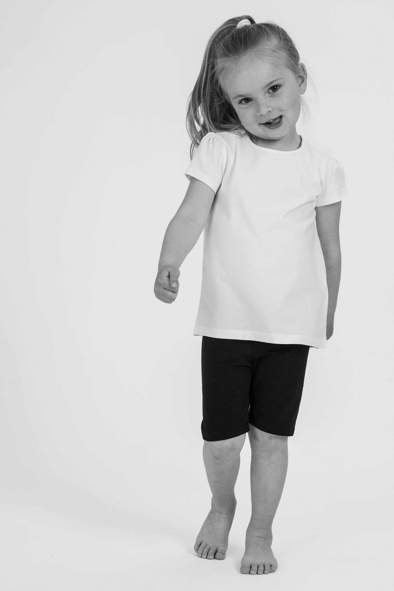 Liten flicka poserar i en vit t-shirt och svarta leggings, med ett leende och en hand uppsträckt i en gest av glädje.