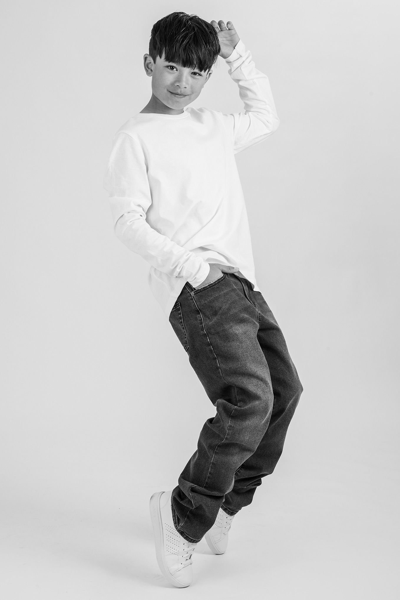 Ung kille i vitt långärmat tröja och mörka jeans poserar lekfullt på en vit bakgrund.