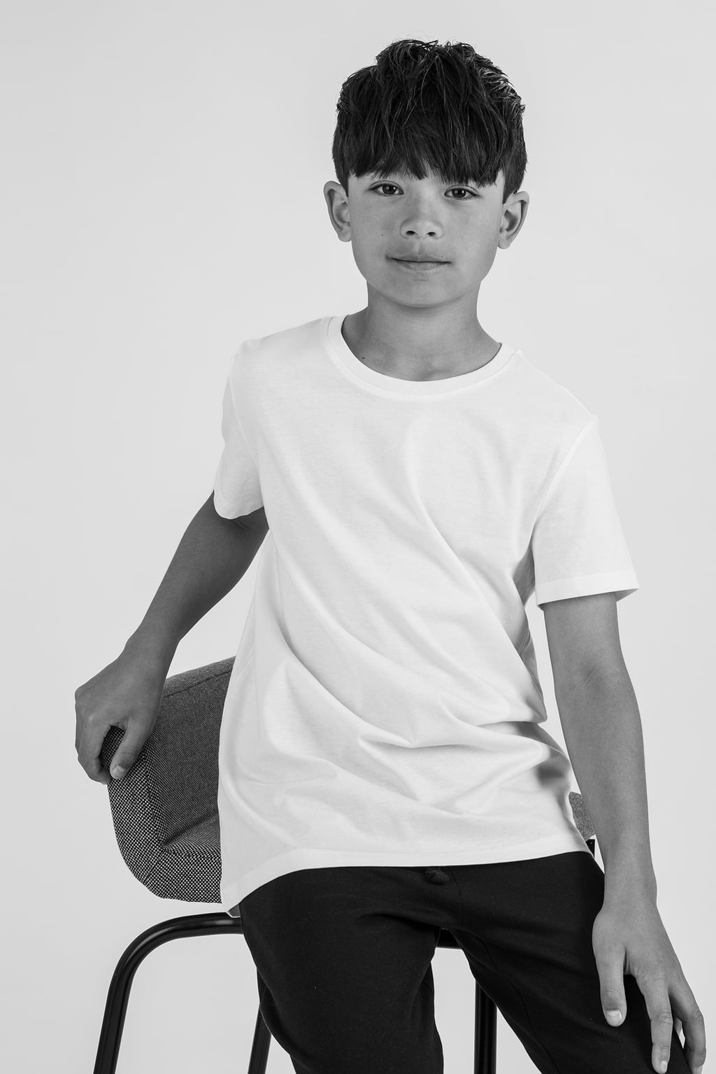 Porträtt av en pojke som sitter på en stol, iförd en vit t-shirt och svarta byxor, med en neutral bakgrund. Bilden fångar en avslappnad och modern stil.