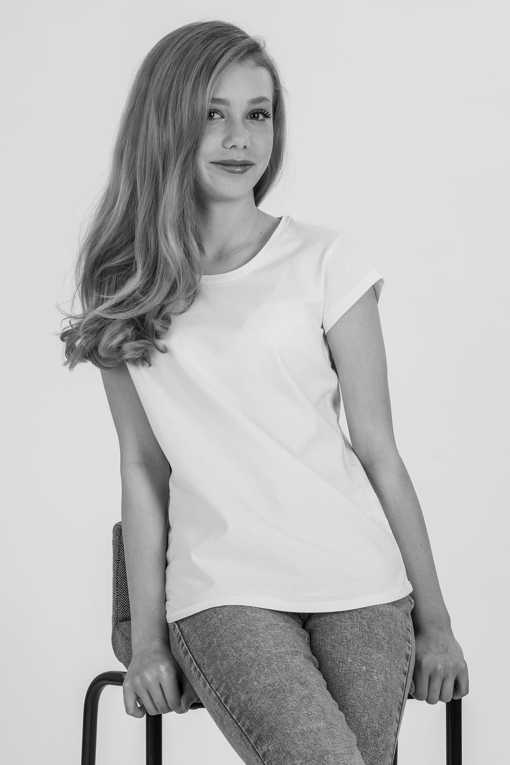 Porträtt av en ung kvinna med långt, lockigt hår, klädd i en vit t-shirt och grå jeans, sittande på en stol mot en neutral bakgrund.
