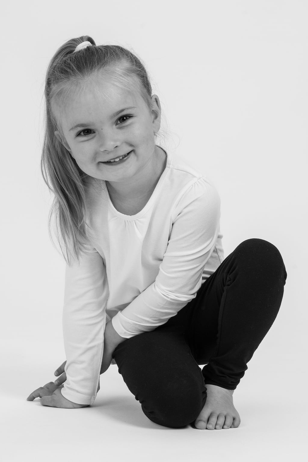 Liten flicka med långt hår i hästsvans, klädd i vit tröja och svarta leggings, sitter på knä och ler mot kameran. Bakgrunden är vit och enkel.