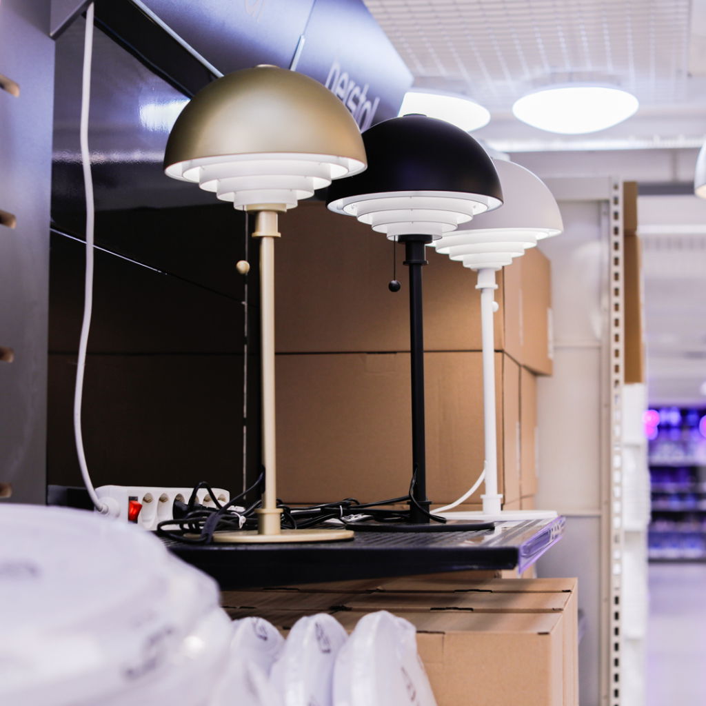Tre moderna bordslampor i olika färger (guld, svart och vit) står på en hylla i en butik. Lamporna har en stilren design med rundad skärm och justerbar ljusstyrka. I bakgrunden syns kartonger och eluttag, vilket ger en känsla av en välsorterad belysningsavdelning.